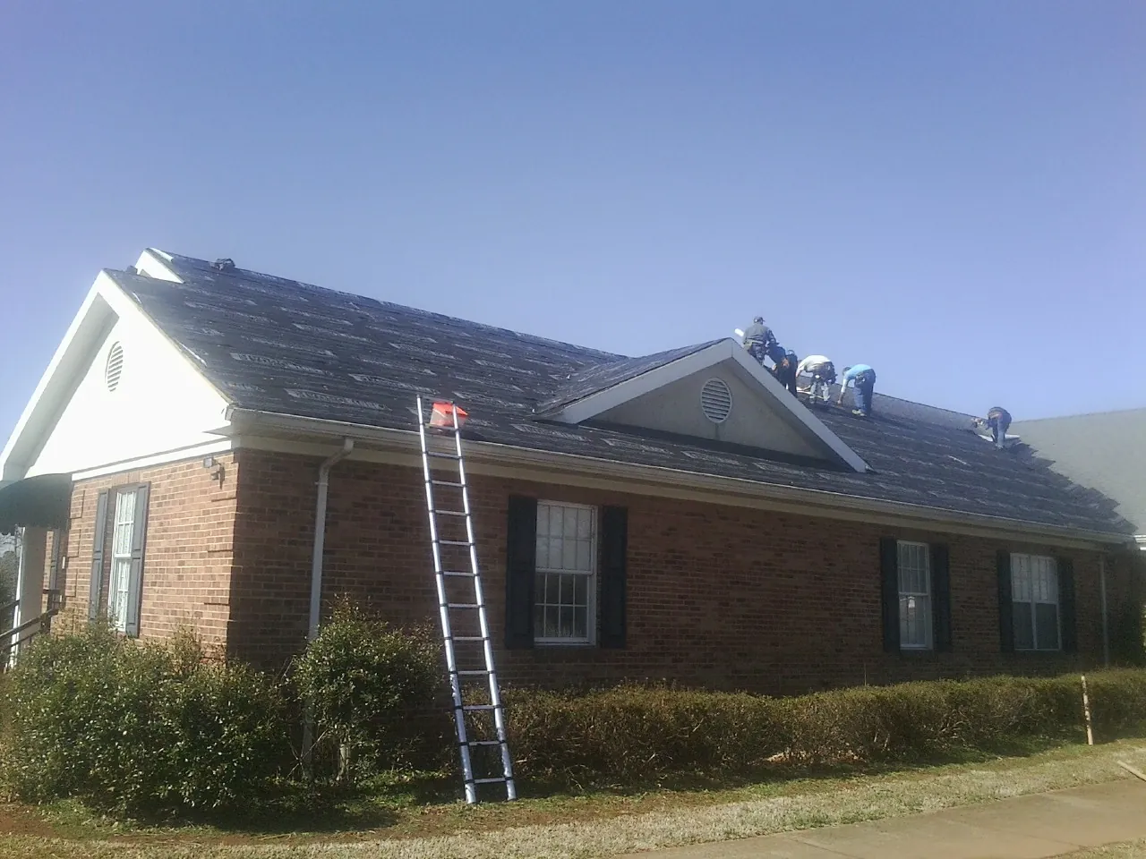 Crew tearing shingles off a brick ranch roof with a ladder set — Emergency Roof Repair work by our crew in Reagan