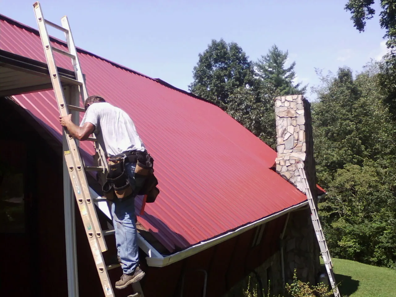Roofer on a ladder beside a red metal roof and stone chimney — Soffit & Fascia Repair by Roofing Brothers in Reagan
